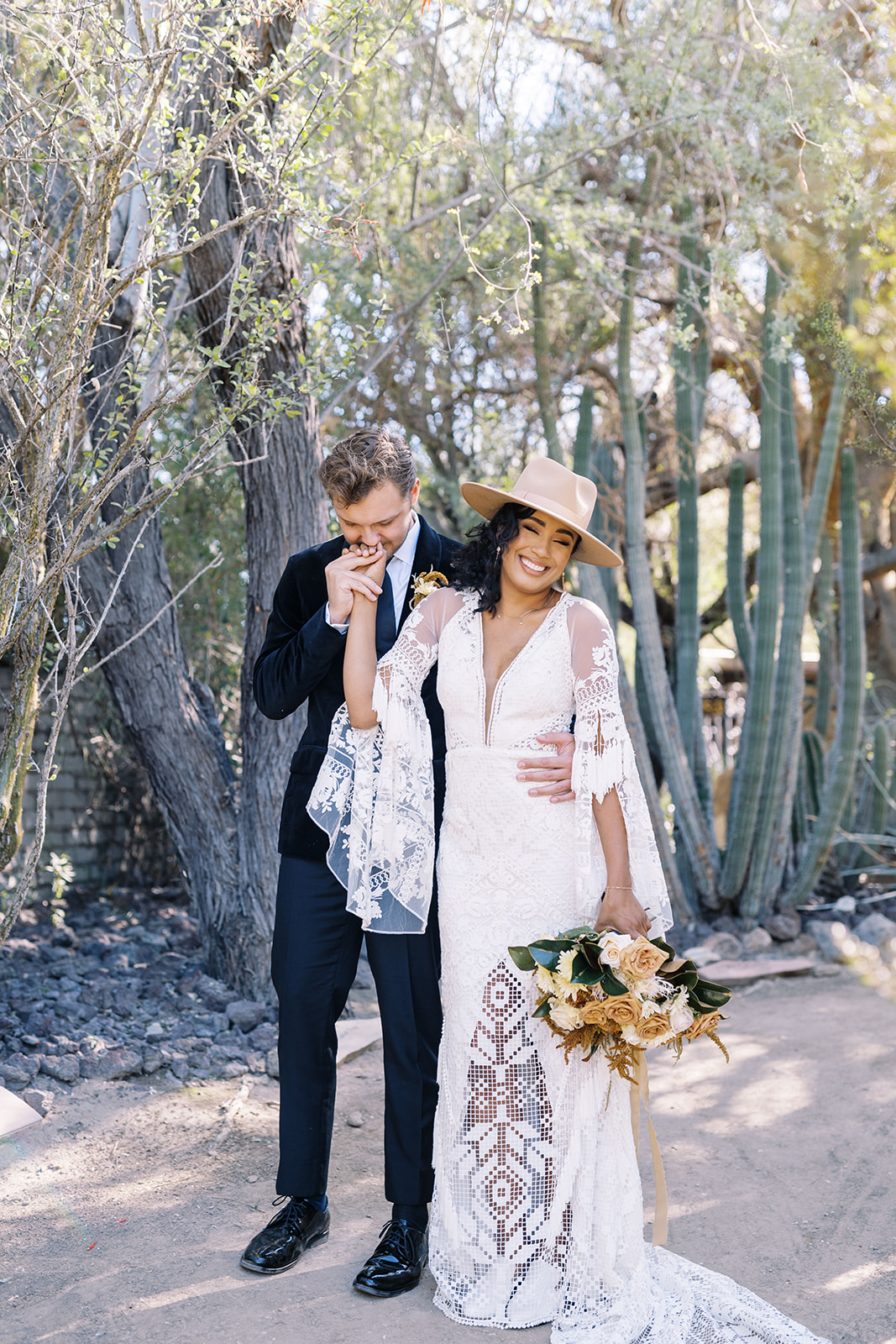Man is kissing woman's hand while standing in the desert surrounded by cacti and trees. Desert elopement planning tips. Elopement photographer in Texas