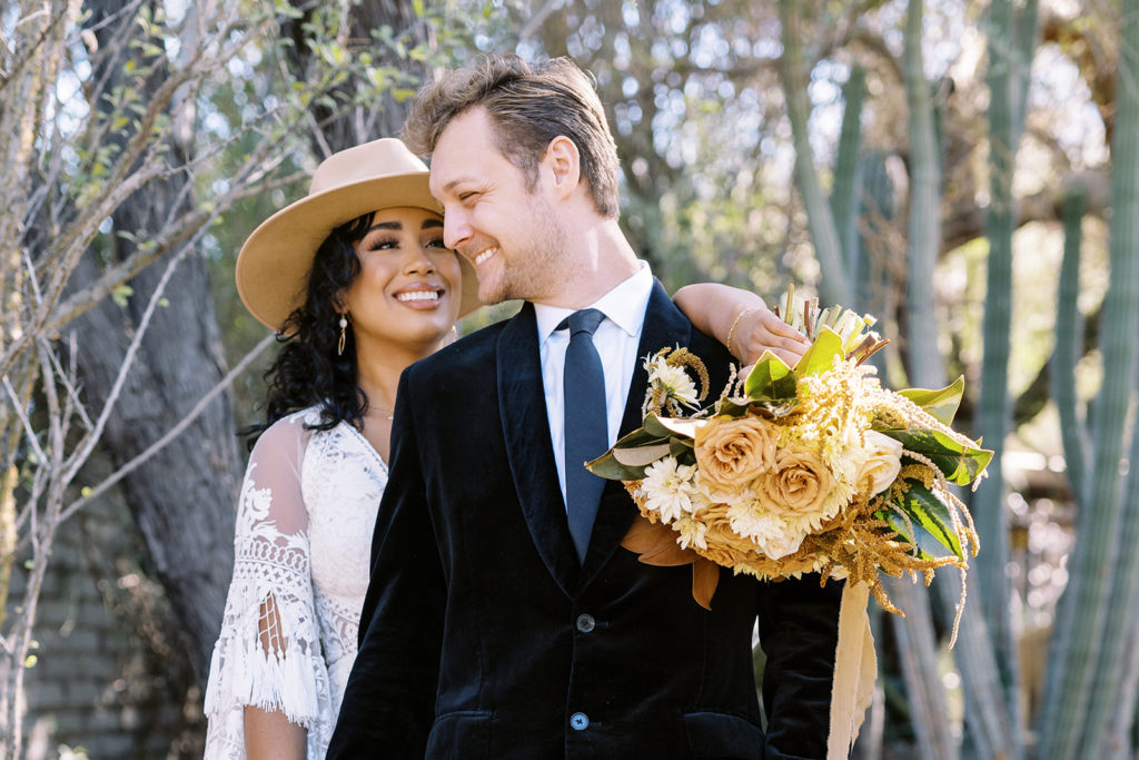Woman smiling while man looks back at her. Woman is holding a wedding bouquet that has natural colored flowers. There is a cactus plant in the background. Texas elopement photographer, desert elopement