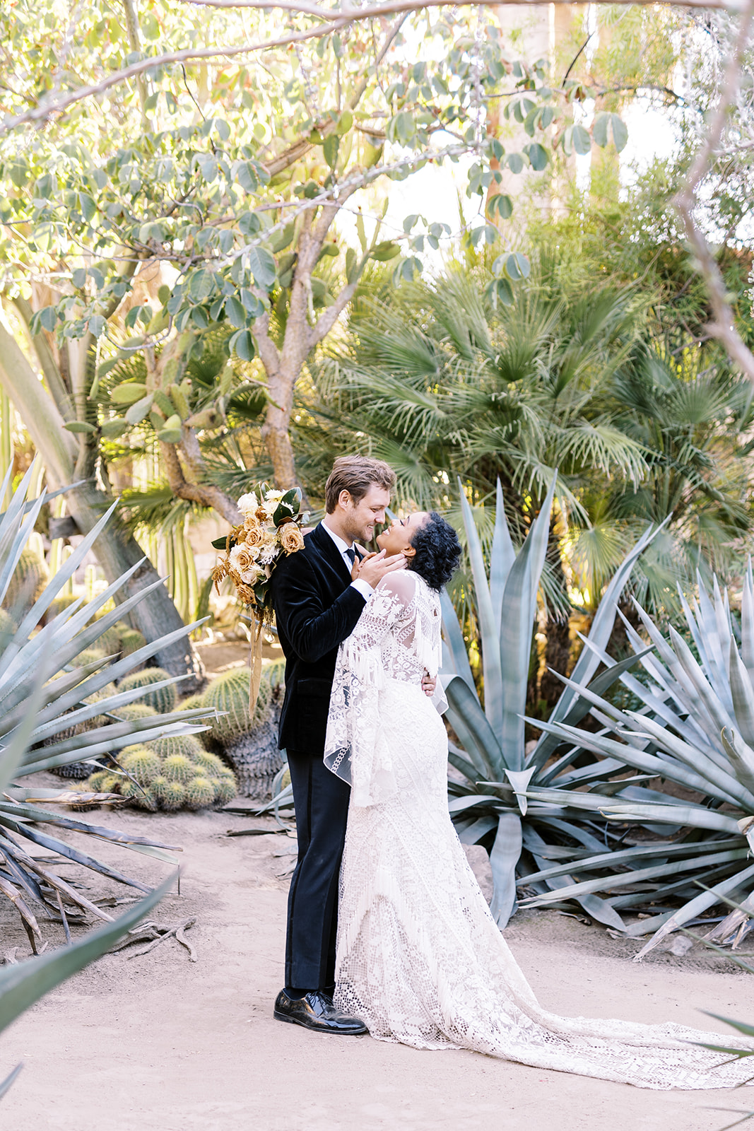 Man and woman hugging while looking into each others eyes while surrounded by desert plants. Woman has curly dark hair while man has light brown colored hair that is medium length. Elopement in the desert. Elopement photographer in Texas