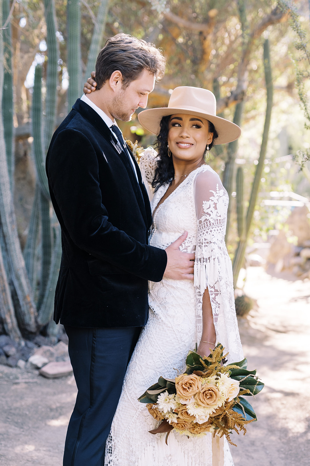 Woman smiling at camera while man looks at her while holding her. They are surrounded by desert cactus and are in a private area. Desert elopement planning