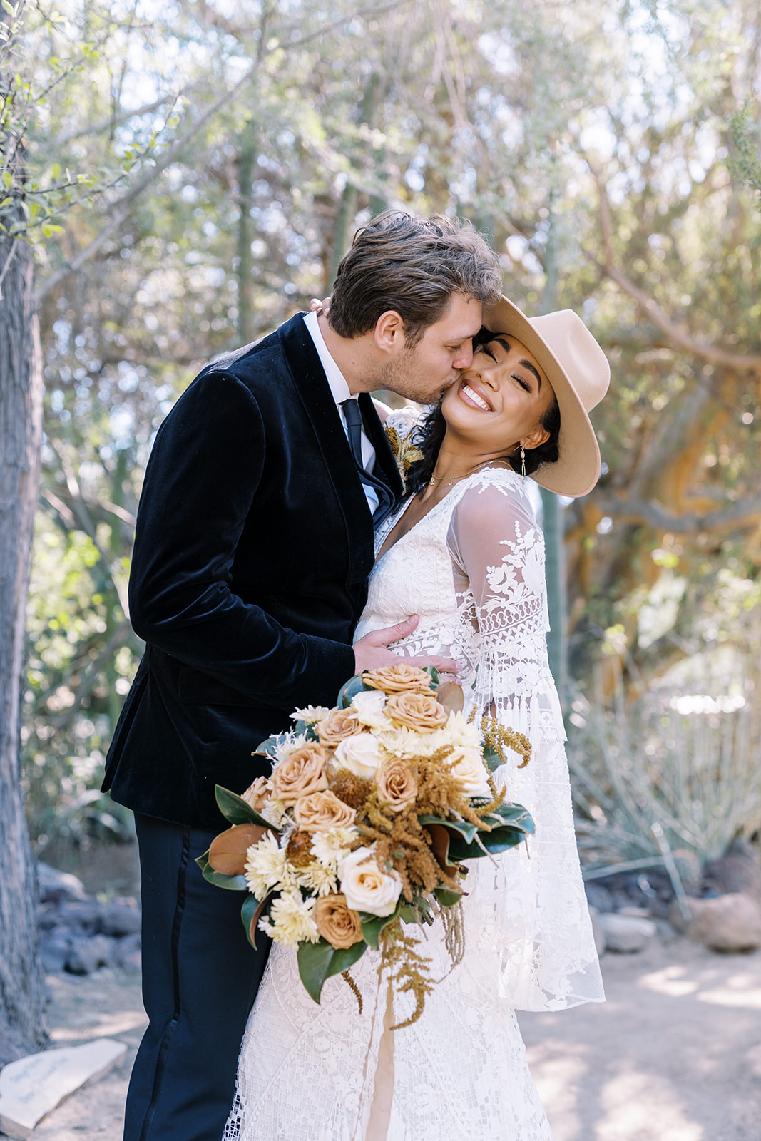 Man kisses woman on cheek while woman smiles. Woman is holding wedding bouquet and is wearing a white lace wedding dress that gives off boho vibes. Desert elopement photos. Elopement photographer in Texas