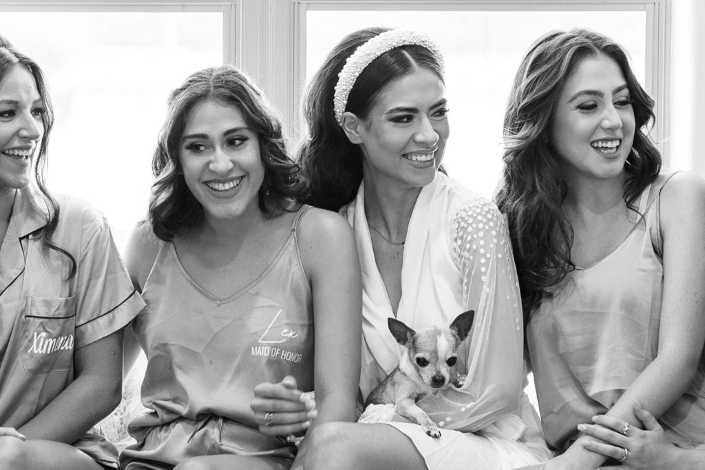 Bridesmaids laughing and sitting with bride who is holding her dog in her lap during bridal portraits. Black and white wedding photos for your wedding day. Texas wedding photographer