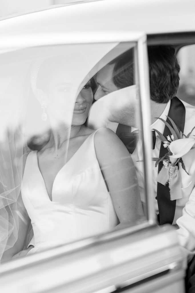 Black and white photo of bride and groom kissing in car with reflection from glass.